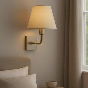 May include: A wall-mounted lamp featuring a brushed brass finish, a square backplate, and a curved arm supporting a conical, off-white fabric lampshade. The lamp emits a warm, soft glow, illuminating the wall behind it with a gentle light.