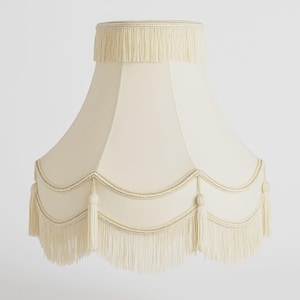 May include: Cream-colored lampshade with a scalloped bottom edge and a fringe trim. The lampshade has a classic, elegant design, with a decorative trim and tassels. The top of the lampshade also features a fringe. The lampshade is made of a soft, fabric material.