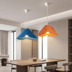 May include: Two pendant lights hanging from the ceiling above a wooden dining table. One light features a blue, petal-shaped shade, while the other has an orange, petal-shaped shade. Both fixtures have white cords and white ceiling mounts.