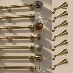May include: A selection of curtain rods with decorative finials in a range of styles, such as a crystal, a speckled orb, and a geometric shape. The rods are a warm brass colour, and the image includes matching curtain hooks.