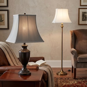 May include: A table lamp with a dark bronze base and a grey square shade sits on a wooden table. A floor lamp with a white shade and a brass base stands nearby. The lamps are in a living room setting with a leather chair and a patterned rug.