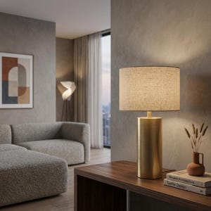 May include: A gold cylindrical table lamp with a beige linen shade sits on a dark wood table. A small terracotta vase with dried plants and two books are next to the lamp. The room has a neutral colour palette with a modern aesthetic.