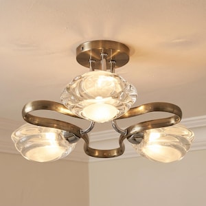 May include: A ceiling light fitting featuring a circular antique brass base with three curved arms extending outwards. Each arm supports a clear, textured glass shade that glows with internal light, creating a balanced and symmetrical design.