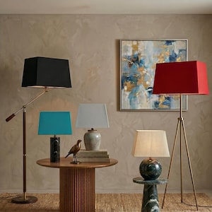 May include: A selection of lamps with rectangular shades in black, teal, grey, and red. The lamps are on a wooden table and a marble side table. A bronze bird sculpture and a painting are also in the room.