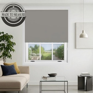 May include: A grey roller blind fitted to a window. The blind is lowered, obscuring the view. The window is white and overlooks a green outdoor scene. The room has a modern design with a sofa and a glass coffee table.