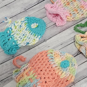 May include: Four crocheted fish-shaped dishcloths in various colours, including pink, blue, green, and orange. The dishcloths are made with a textured stitch and have a small crocheted circle in the centre of each fish.