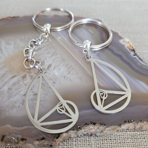 May include: A pair of silver keychains with a geometric design. Each keychain features a circle with a triangle inside, and a smaller circle inside the triangle. The keychains are connected by a silver chain.