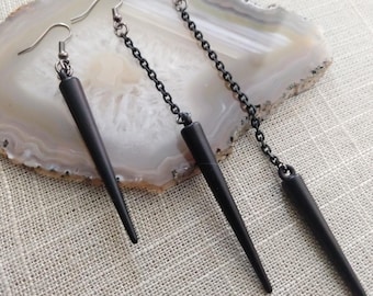 Pode incluir: Brincos pretos em forma de ponta, com corrente e fecho de gancho. Os brincos são exibidos numa superfície clara e estampada. Têm um acabamento mate escuro e uma forma longa e cónica.