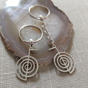 May include: Two silver-colored keychains with a Reiki symbol pendant. The pendants feature a spiral design within a rectangular shape. One keychain has a chain link, while the other has a simple ring.