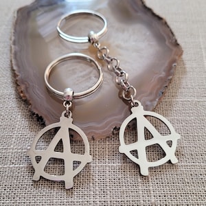 Peut inclure: Deux porte-clés argentés avec un charme symbole d'anarchie circulaire. Les porte-clés sont attachés à des anneaux de clés argentés.