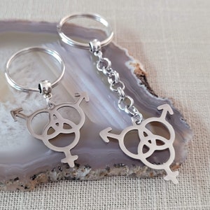 May include: Two silver keychains with a transgender symbol pendant. The pendants feature a stylized combination of male, female, and arrow symbols. The keychains are made of a shiny metal and are displayed on a light-colored surface.