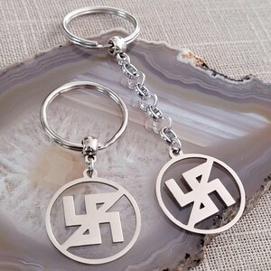 Peut inclure: Deux porte-clés argentés avec un pendentif circulaire représentant un symbole swastika stylisé. Les porte-clés sont attachés à des anneaux de clés argentés.