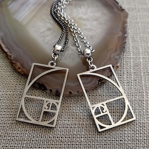 May include: Two silver necklaces with a geometric design pendant featuring the Fibonacci spiral. The necklaces are on a light brown textured surface.