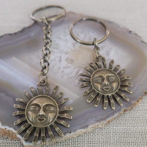 Könnte beinhalten: Zwei antike silberfarbene Schlüsselanhänger mit einem Sonnengesicht-Design. Einer hat eine Kette, der andere einen einfachen Ring. Die Sonne hat ein lächelndes Gesicht mit Strahlen, die sich ausbreiten.