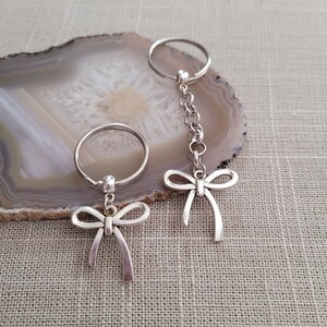 May include: Two silver keychains with bow-shaped charms. The keychains are on a gray and white agate stone background.
