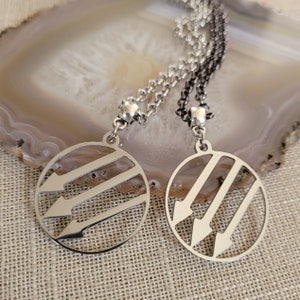 May include: Two silver necklaces with circular pendants featuring a stylized three-arrow design. The necklaces are on a light brown background.