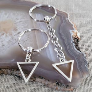 Może przedstawiać: Dwa srebrne breloki z trójkątnymi zawieszkami. Breloki są połączone srebrnym łańcuszkiem.