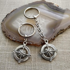 May include: Two silver compass keychains with a chain connecting them. The compass features a star design with the letters N, S, E, and W around the edge.