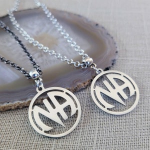 Könnte beinhalten: Zwei silberne Halsketten mit einem runden Anhänger, der die Buchstaben "NA" in einem stilisierten Design zeigt. Die Halsketten liegen auf einer strukturierten Oberfläche.