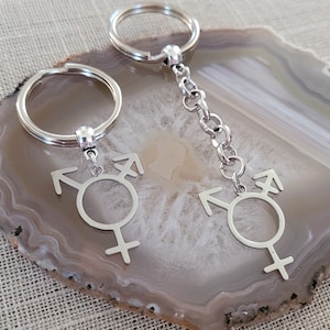 May include: Two silver keychains with a gender symbol charm. The charms are silver and feature the transgender symbol.