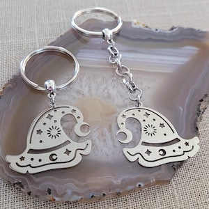 May include: Two silver keychains with a witch hat design. The hats have a crescent moon, stars, and a sun design. The keychains are on a gray and white agate background.