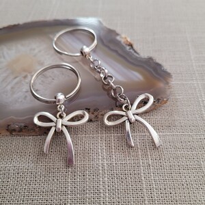 May include: Two silver keychains with bow charms. The keychains are attached to silver rings. The rings are on a light brown agate surface.