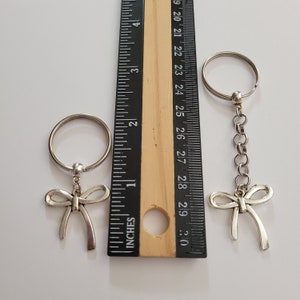 May include: Two silver keychains with a bow design. The keychain on the left is attached to a silver key ring. The keychain on the right is attached to a silver chain and a silver key ring. Both keychains are measured against a ruler.