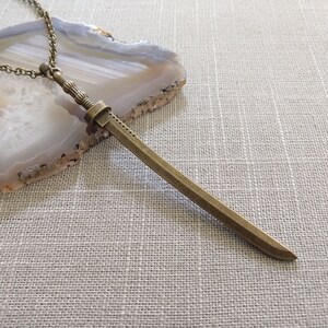May include: A bronze-toned metal sword pendant necklace featuring a detailed hilt with ribbed textures and a straight blade. The pendant is attached to a matching bronze-colored link chain, showcasing a metallic finish and a slender design.