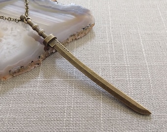 Pode incluir: Um colar com pingente de espada em metal tom bronze, apresentando um punho detalhado com texturas caneladas e uma lâmina reta. O pingente está preso a uma corrente de elos correspondente, exibindo um acabamento metálico e design fino.