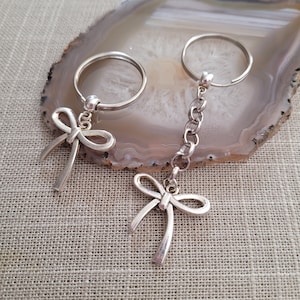 May include: Two silver keychains with bow charms. One keychain has a single bow charm, while the other has a bow charm attached to a silver chain.