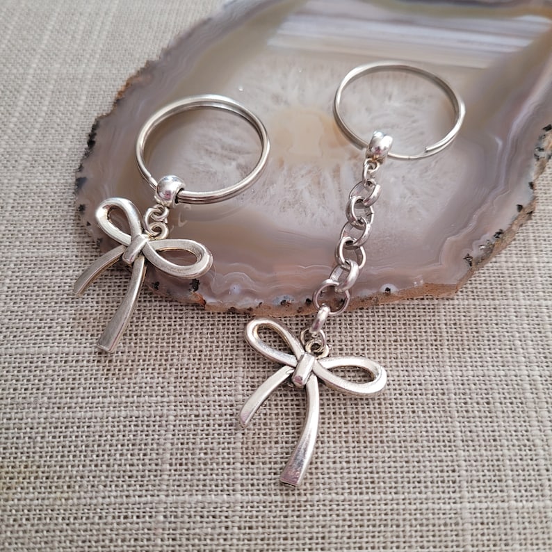 May include: Two silver keychains with bow charms. One keychain has a single bow charm, while the other has a bow charm attached to a silver chain.