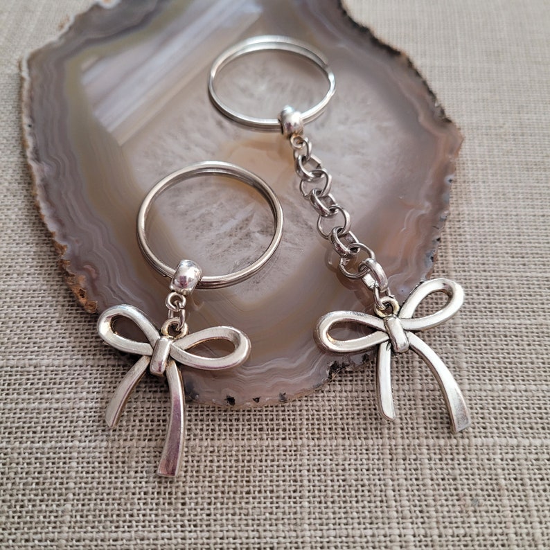 May include: Two silver keychains with bow-shaped charms. The keychains are on a gray and white agate stone background.