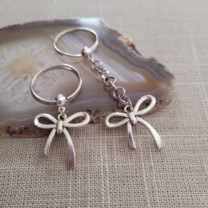 May include: Two silver keychains with bow charms. The keychains are attached to silver rings. The rings are on a light brown agate surface.