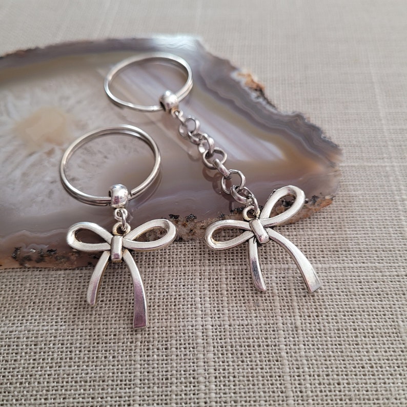 May include: Two silver keychains with bow charms. The keychains are attached to silver rings. The rings are on a light brown agate surface.