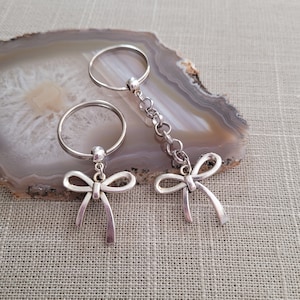 May include: Two silver keychains with bow charms. The keychains are on a light brown agate surface.