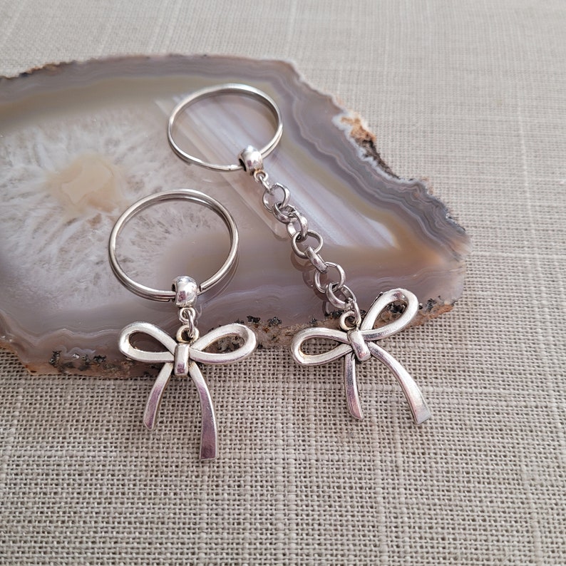 May include: Two silver keychains with bow charms. The keychains are on a light brown agate surface.