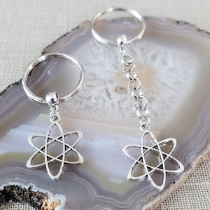 May include: Two silver keyrings featuring atom-shaped pendants. One keyring has a short chain, the other a simple ring. The pendants are six-pointed star designs. The keyrings are displayed on a grey and white stone surface.