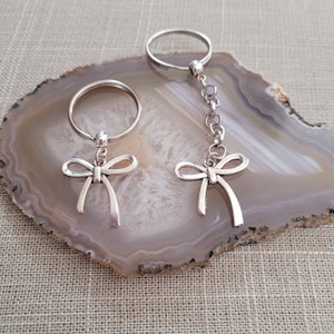May include: Two silver keychains with bow charms. One keychain has a silver ring and the other has a silver chain.