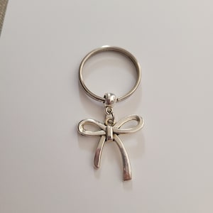 May include: A silver key chain with a bow charm. The key chain is made of metal and has a silver finish. The bow charm is attached to the key chain with a silver ring.