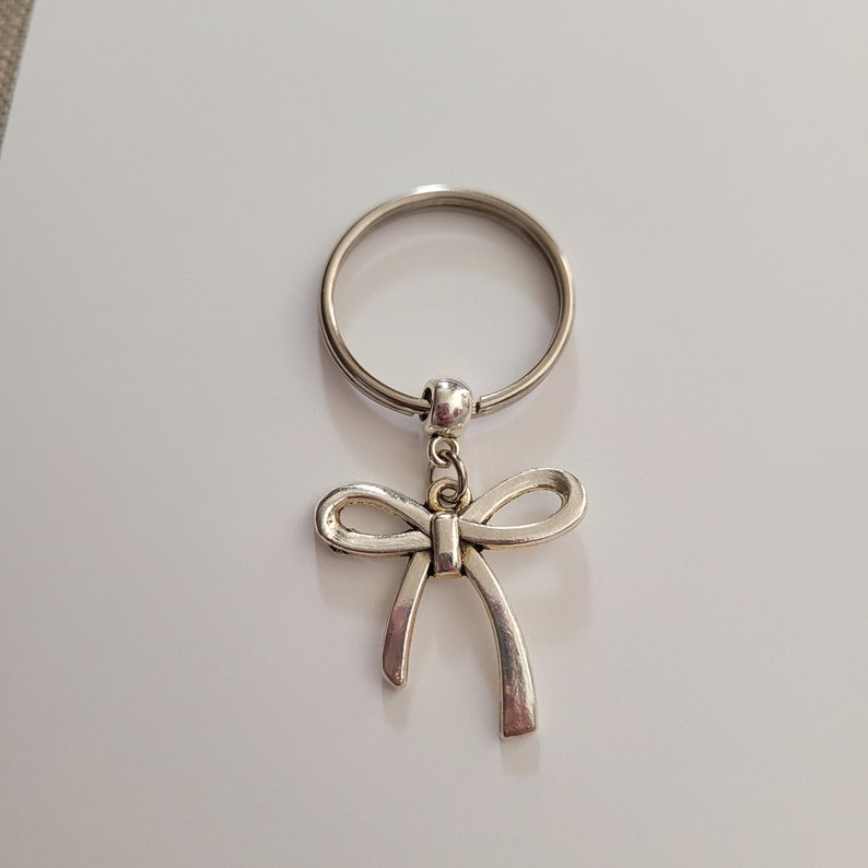 May include: A silver key chain with a bow charm. The key chain is made of metal and has a silver finish. The bow charm is attached to the key chain with a silver ring.