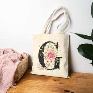 Op de afbeelding: Een crèmekleurige draagtas met een grote zwarte letter "G" versierd met een aquarel bloemenontwerp. De bloemenarrangement bevat een grote roze roos, kleinere roze bloemen en groene bladeren. De tas heeft crèmekleurige handvatten.