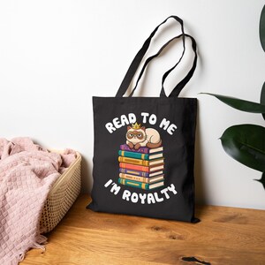 Op de afbeelding: Zwarte draagtas met een cartoonkat met een kroon, zittend op een stapel kleurrijke boeken. De tekst "Read to me I'm Royalty" is in het wit gedrukt. De tas heeft zwarte handvatten.