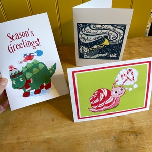 May include: Three Christmas cards with different designs. The first card features a green dinosaur wearing a red scarf and a white hat with a red pom-pom. The second card features a blue and white illustration of a person playing a trumpet with the text "Even when the world feels sad I will make a joyful noise". The third card features a pink snail with a candy cane shell and a thought bubble with a candy cane inside.