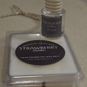 May include: A clear glass car diffuser bottle with a light wood cap and a black label reading Moondog Creations Strawberry Leather Diffuser, paired with a square white soy wax melt in a plastic container with a black label reading Strawberry Leather.