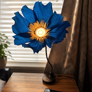 Könnte beinhalten: Eine blaue, blütenförmige Tischlampe mit einem hellgelben Zentrum. Die Lampe steht auf einem Holztisch mit schwarzem Sockel und gebogenem Stiel. Ein Smartphone wird auf dem Tisch aufgeladen.
