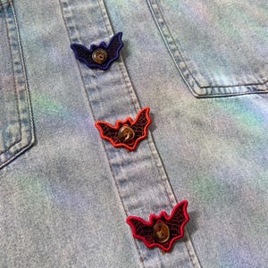 May include: A light blue denim shirt featuring a tie-dye pattern in shades of green and purple. The shirt has a button-down front with four bat-shaped embroidered patches in pink, blue, red, and dark pink, plus one on the chest pocket.