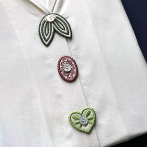 Può includere: Primo piano di un capo bianco con decorazioni ricamate decorative. Include un design a forma di foglia, un ovale e un cuore, ciascuno con un bottone al centro. Il ricamo utilizza tonalità verdi e rosse.