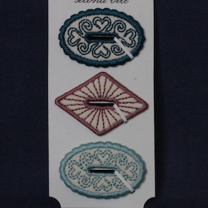 Può includere: Tre abbellimenti in tessuto ricamato su una carta bianca. Quelli superiori e inferiori sono di forma ovale con ricami verde acqua e azzurri, con disegni a cuore e a spirale. Quello centrale è a forma di diamante con ricamo rosa. La carta recita "second bite".