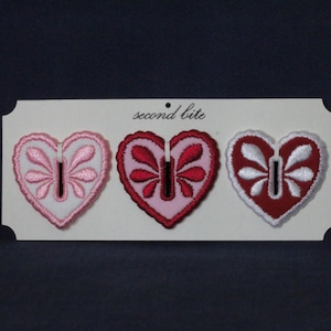 Può includere: Tre toppe ricamate a forma di cuore in rosa, rosso e bianco, ciascuna con un design a forma di buco della serratura e accenti floreali. Le toppe sono esposte su una carta bianca con la scritta "second bite" in alto.