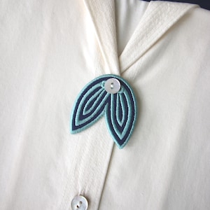 Può includere: Spilla ricamata a forma di foglia, blu acqua e blu navy, con un bottone bianco al centro. La spilla è attaccata a una camicia color crema, con un secondo bottone visibile sotto.